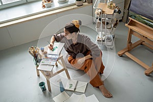 Top view of focused woman artist sitting on floor in art studio painting watercolours in sketchbook photo