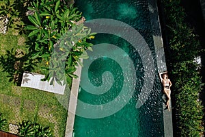 Top view of woman in beige bikini relaxing on edgeof infinity pool around palm trees and jungle. Luxury holiday.Girl