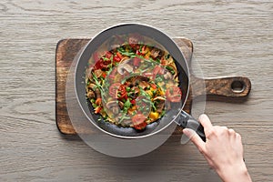 Top view of woman cooking omelet with mushrooms, tomatoes and greens on frying pan on wooden board.