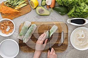 Top view of woman cutting with knife spring rolls on cutting board.