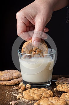 Top view of woman hand with chocolate chip cookie in glass of milk, on wooden table with cookies and crumbs, selective focus, with