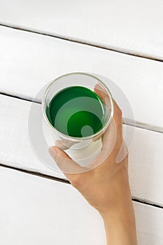 Top view of woman hand holding cup of chlorophyll drink on white wooden background. Vertical image