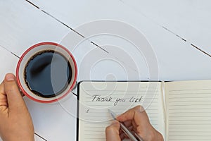 Top view of woman hand writing thank you list and holding cup of coffee on white wooden table
