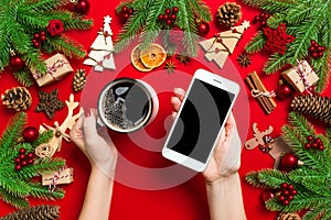 Top view of a woman holding a phone in one hand and a cup of coffee in another hand on red background. Christmas decorations and