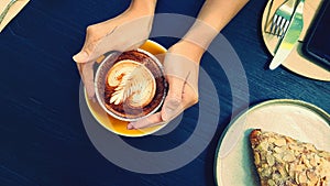 Top view of woman`s hand holding art latte coffee with almond cream croissant, knife, fork and phone on blue wooden background