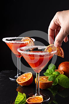 Top view of woman`s hand holding half slice of red orange over martini glass, on black slate with red half orange and mint, black