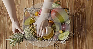 Top view woman`s hand puts natural organic mango, pineapple, kiwi in a the box with variety exotic tropic fruis on a