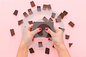 Top view on woman`s hands breaking a piece of dark chocolate above pink table background