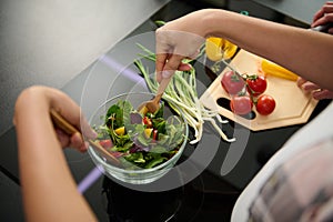 Top view of woman`s hands holding wooden spoons and mixing ingredients in a glass bowl, preparing delicious healthy salad for