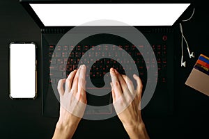 Top view woman`s hands typing on laptop  with white blank screen. Empty screen smartphone, headphones, credit cards on black