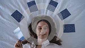 Top view. A woman in a white shirt lies on a white background surrounded by tarot cards and covers her face with the