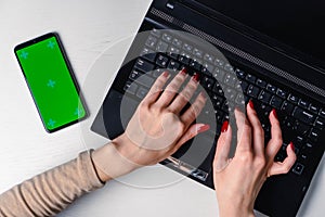 Top view of woman& x27;s hands typing on laptop keypad placed on white office desktop with blank smartphone