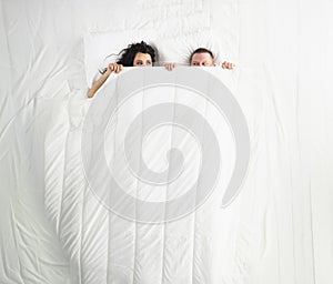 Woman and man peeking on each other under white blanket, awake couple in bed