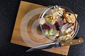 Top view of wooden cutting board, knife and glass bowl full of vegetables scraps on the kitchen table.Empty space