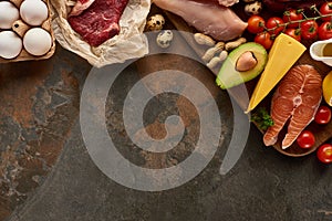 Top view of wooden cutting board with raw fish, meat, poultry, cheese, eggs, vegetables and peanuts on dark brown marble