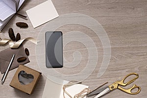 Top view of wooden desk and phone with empty screen,present box,ribbon,cones,cards,scissors on it.Christmas gifts buying and wrapp