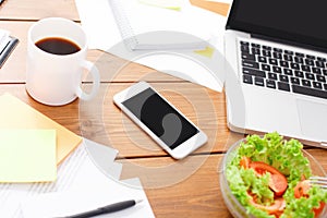Top view of wooden desk with salad, laptop, smartphone and other items