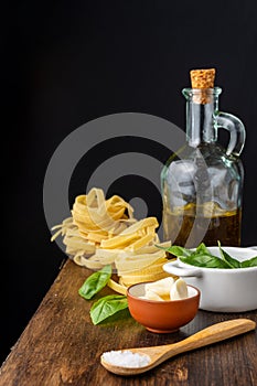 Top view of wooden spoon with salt, cut garlic, basil, bottle with olive oil and raw tagliatelle, on wooden table, black backgroun