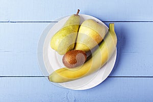 Top view on wooden table with heap fresh fruit. Pears, banana and kiwi fruit on white plate. Copy space