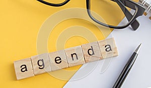 Top view the word of the agenda written on wooden cubes on the desktop with glasses  pen  paper Notepad on a yellow background