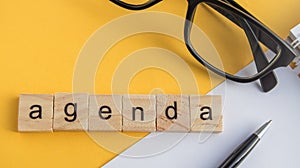 Top view the word of the agenda written on wooden cubes on the desktop with glasses, pen, paper Notepad on a yellow background,