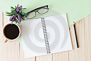 Top view of working desk with blank notebook with pencil, coffee cup, eyeglasses and plant on wooden background