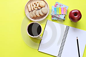 Top view of working desk with blank notebook with pencil, cookies, apple, coffee cup and colorful note pad on yellow background