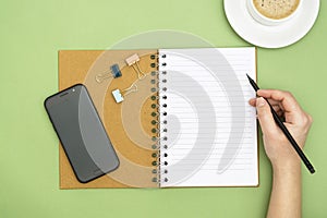 Top view of working table. Open notebook, coffee cup, smartphone and a woman hand holding an pencil, writting a message. Copy