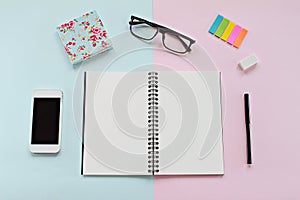 Top view of workspace desk with blank notebook, eye glasses, smart phone and office stationery on pink and blue background