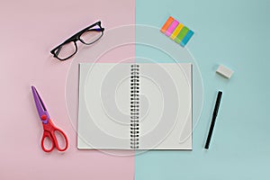 Top view of workspace desk with blank notebook, glasses and office stationery set on pink and blue background
