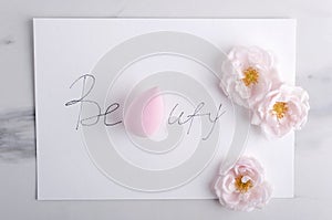 Top view of written wird Beauty, pink cosmetic sponge and roses on the marble surface