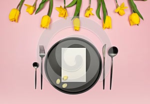 Top view of yellow tulips, daffodils, easter eggs on pink background. Table setting, black plate and cutlery, white card.