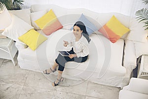 Top view of Young beautiful Asian woman look at the camera sitting on white sofa using tablet smartphone explore website. reading