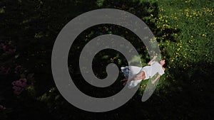 Top view of a young beautiful woman in a white dress with dark hair lying on the green grass with yellow flowers.