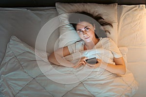 Top view of young Caucasian woman using smartphone and looking at camera lying in bed at night. Insomnia and social