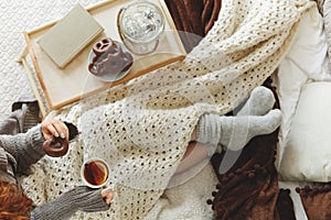 Top view of young girl covered with cozy white blanket siting on bed drinking tea and eating