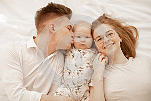 Top view of young happy family in white clothes with plump cherubic baby infant toddler lying on white cotton bed.