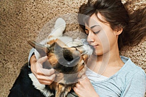top view young joyful woman lying on carpet with cute pet. Happy girl kissing and stroking Welsh Corgi dog.