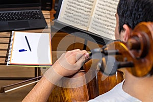 Top view of a young man playing the cello in the middle of an online class with a laptop computer and a notebook on the table with