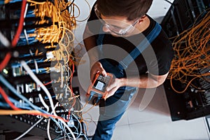 Top view of young man in uniform with measuring device that works with internet equipment and wires in server room