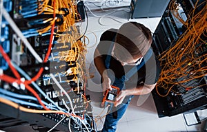 Top view of young man in uniform with measuring device that works with internet equipment and wires in server room