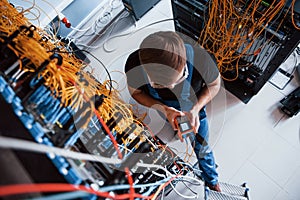Top view of young man in uniform with measuring device that works with internet equipment and wires in server room