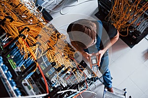 Top view of young man in uniform with measuring device that works with internet equipment and wires in server room