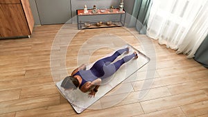Top view of young woman in bodysuit exercising, doing Four Limbed Staff yoga pose on a mat in living room at home on a