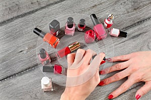 Top view - young woman hands, applying second layer of nail varnish, with more bottles of polish in back, on gray wood desk. All
