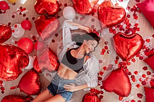 Top view young woman lying on wooden floor surrounded by red heart shaped balloons in Valentines Day