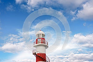 Top of a white-red lighthouse against a blue sky with an airplane