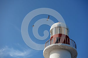 Top of a white-red lighthouse against a blue sky with an airplane.