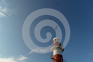 Top of a white-red lighthouse against a blue sky with an airplane.