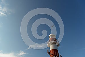 Top of a white-red lighthouse against a blue sky with an airplane.